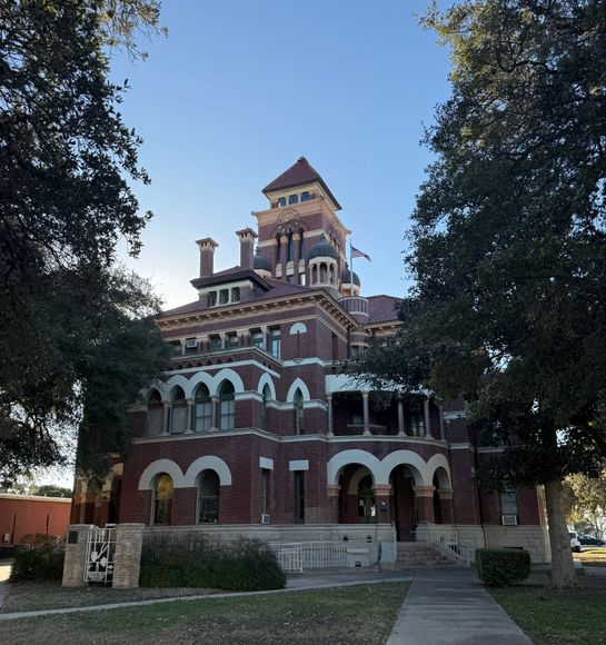An ornate, multi-story brick courthouse with arches and a central tower, framed by large trees under a clear blue sky.