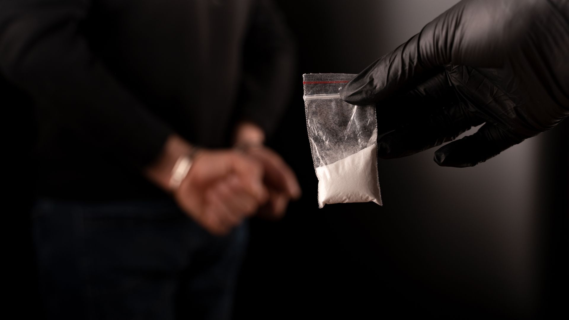 A person in a black glove holds a small plastic bag filled with a white powder in front of another person.