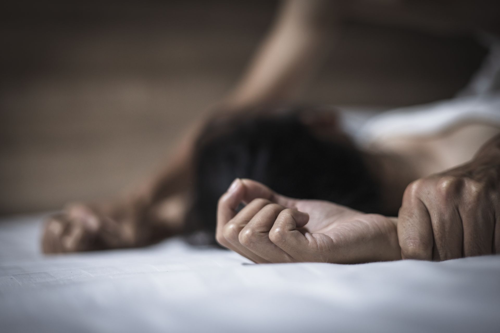 Close-up of hands held tightly together on a white sheet, with a person's head resting in the background.