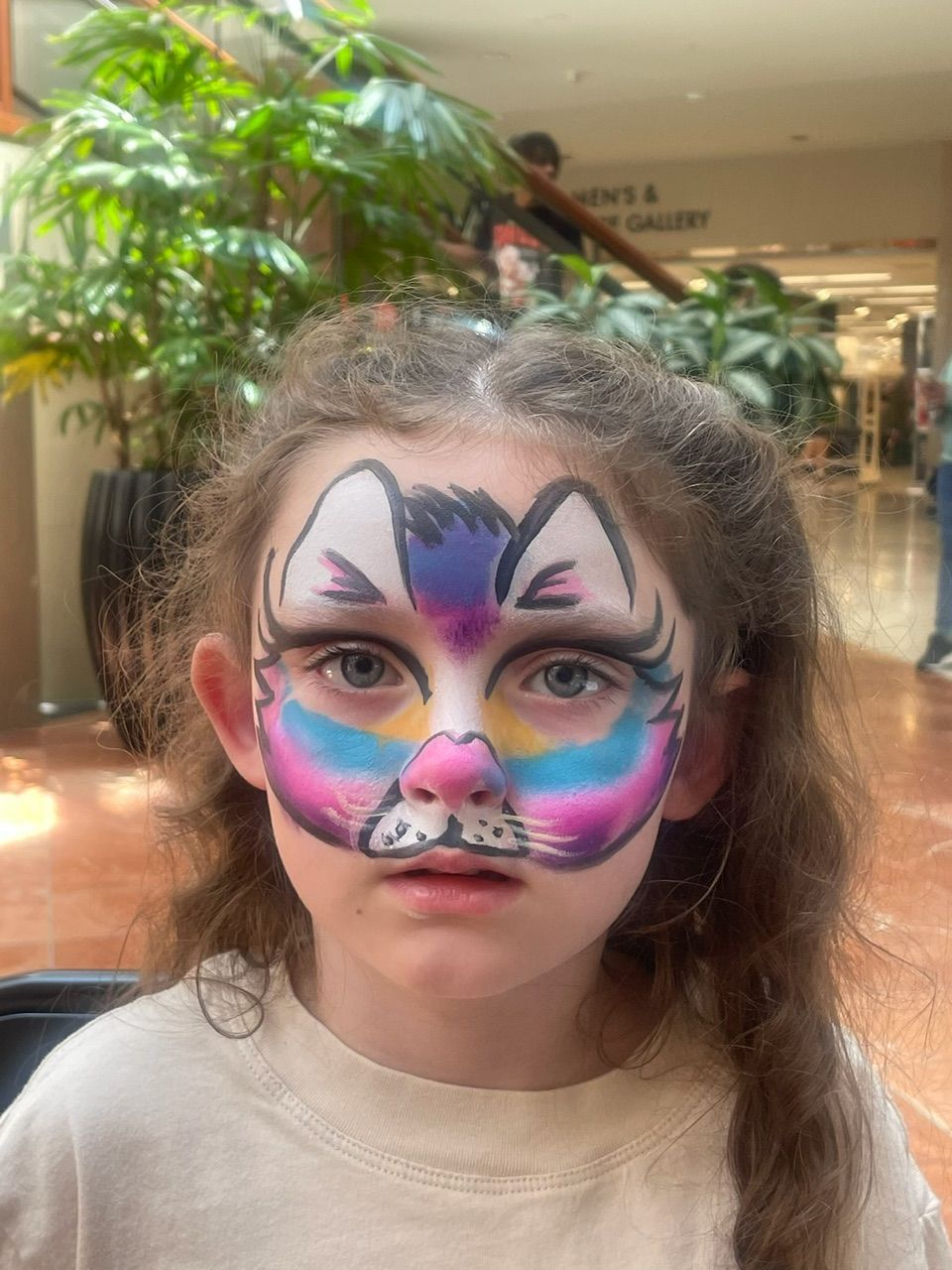 Child with colorful cat face paint in an indoor mall, looking at the camera.