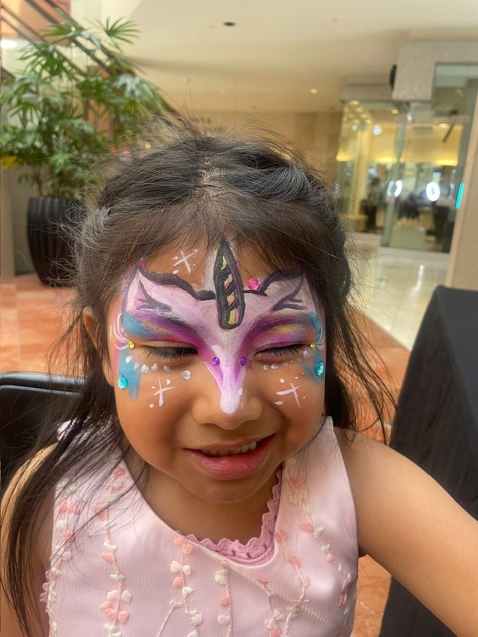 Child with colorful butterfly face paint smiling indoors near a mall seating area