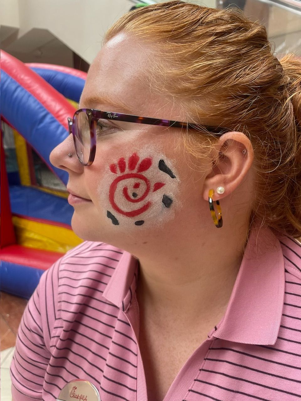 Person in glasses with a red-and-black face paint design, wearing a pink striped shirt, looking left.