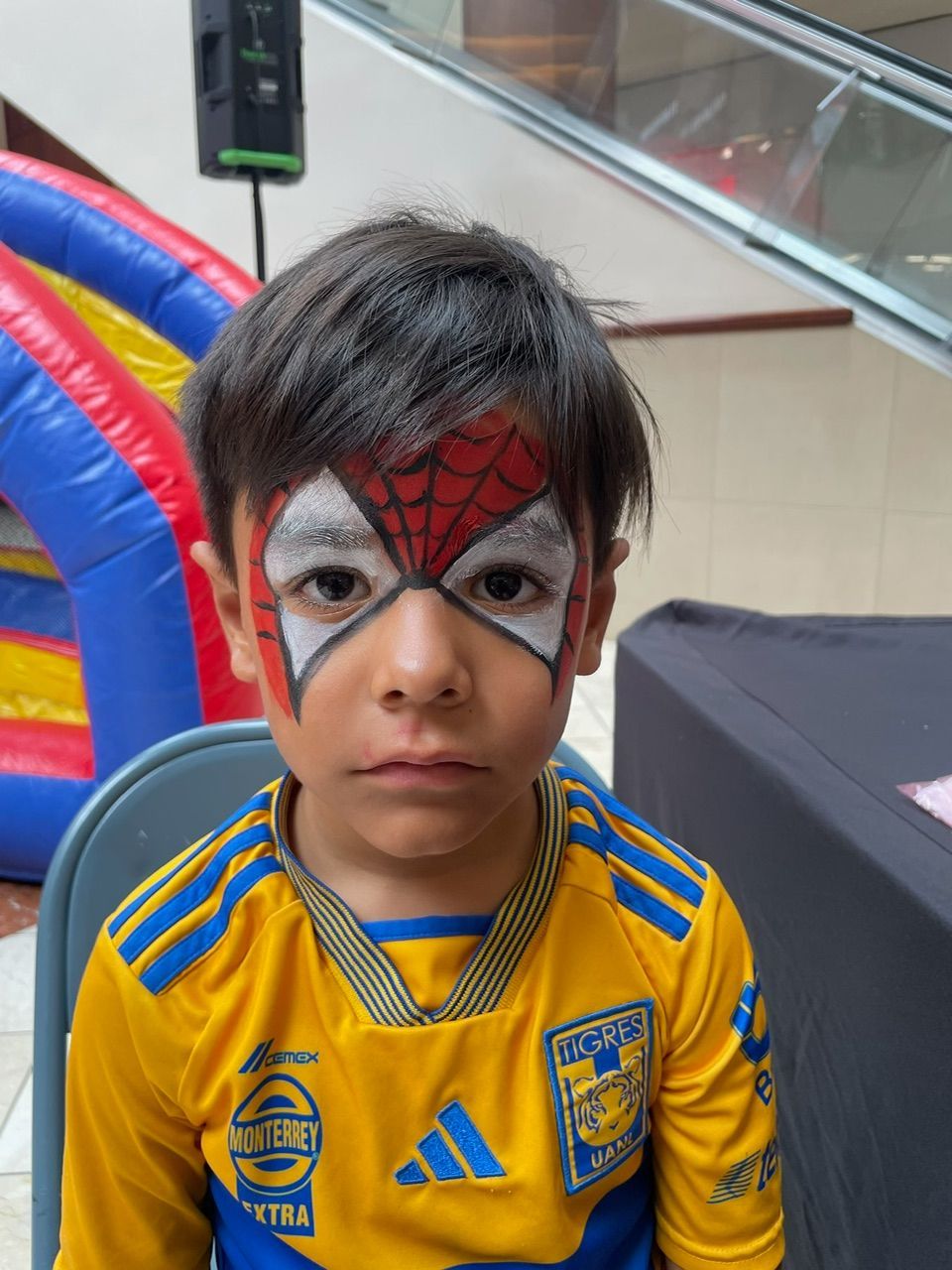 Child in a yellow soccer jersey and red-and-silver face paint looks at the camera indoors.