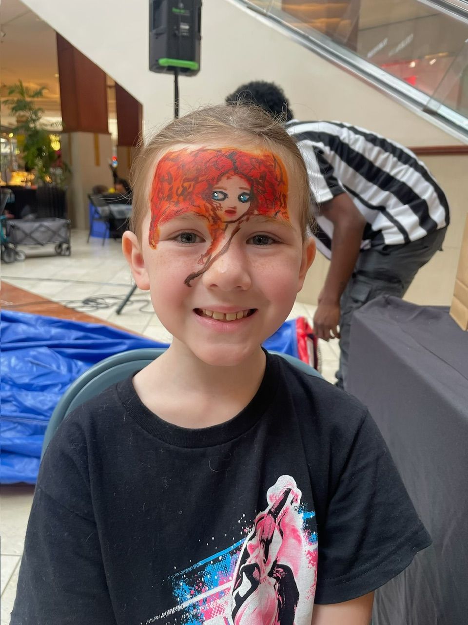 Smiling child with painted face in a mall, with a person in a striped shirt behind them