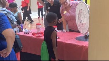 People at a pink-covered fair booth with a child facing a vendor and a large white fan on display