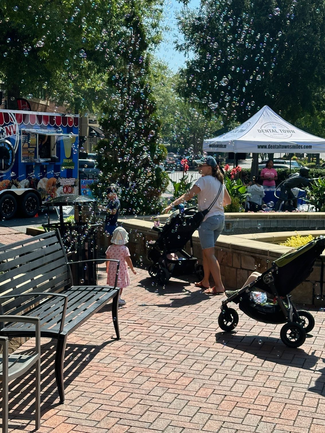 Person pushing a stroller in a sunny park with benches, trees, and a white tent
