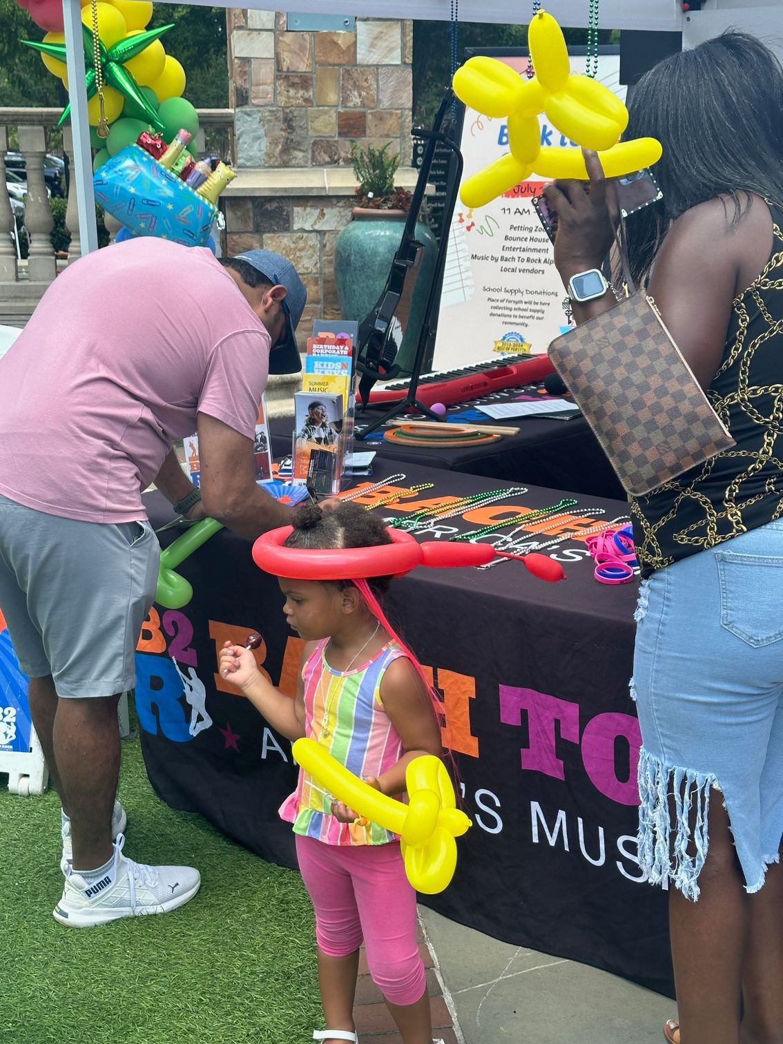 People at a balloon-decorated table, with a child holding yellow balloon animals outdoors.