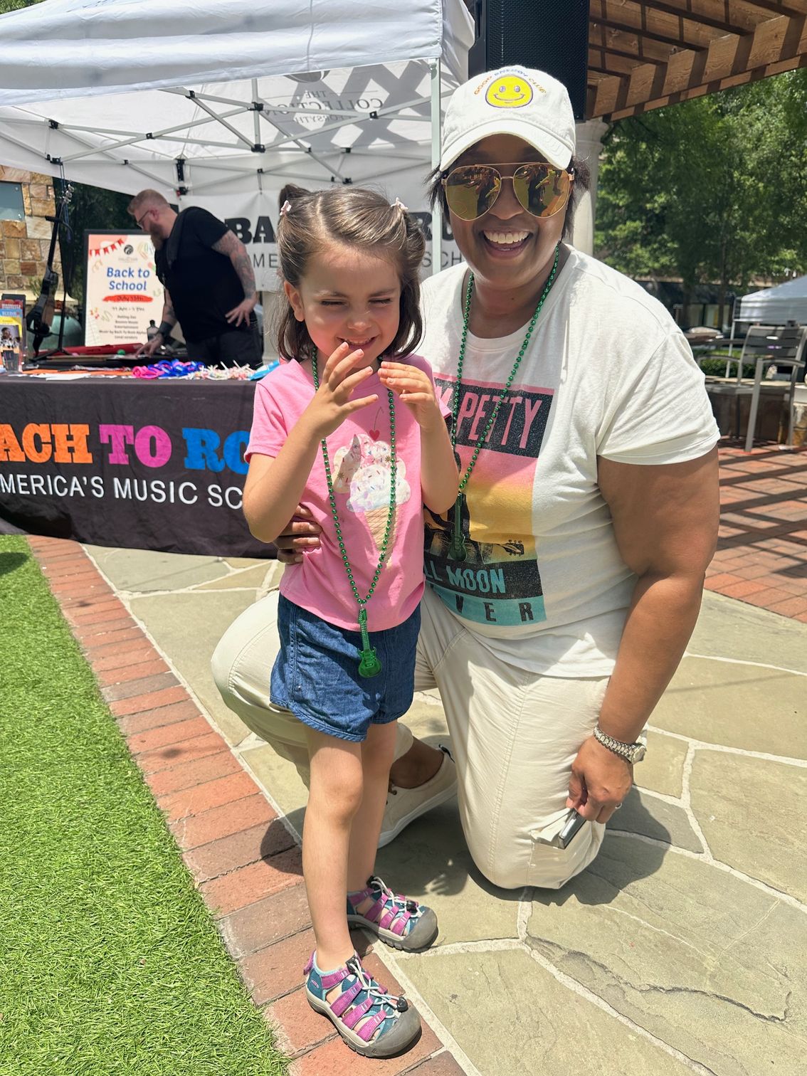 Two people posing at an outdoor music festival booth, one kneeling beside a child holding a snack.