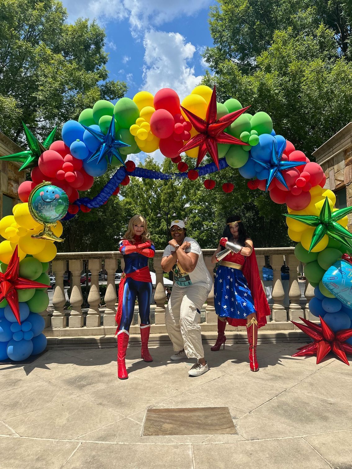 Three costumed performers posing under a colorful balloon arch outdoors