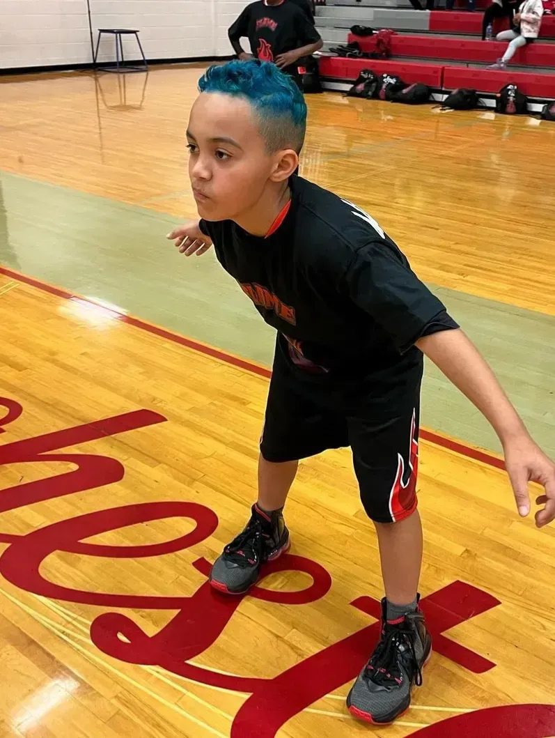 Child in a black T-shirt and shorts crouches on a gym court with blue hair, ready to move.