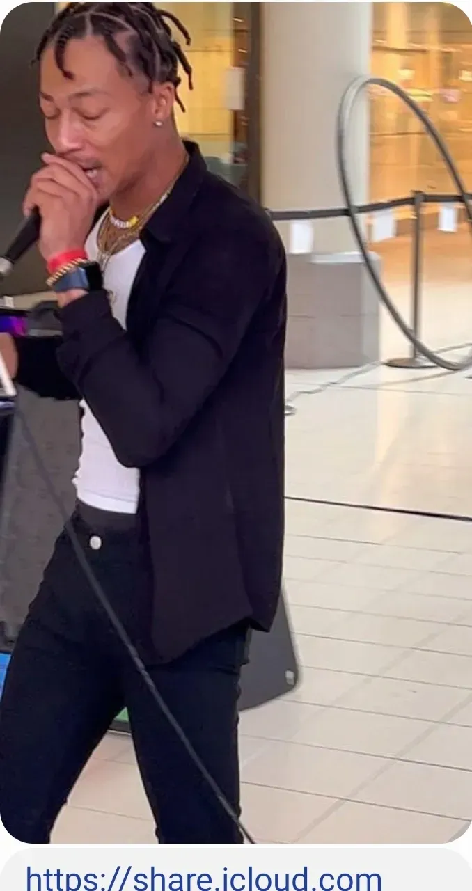 Person singing into a microphone in a bright indoor space, wearing a black blazer and white shirt