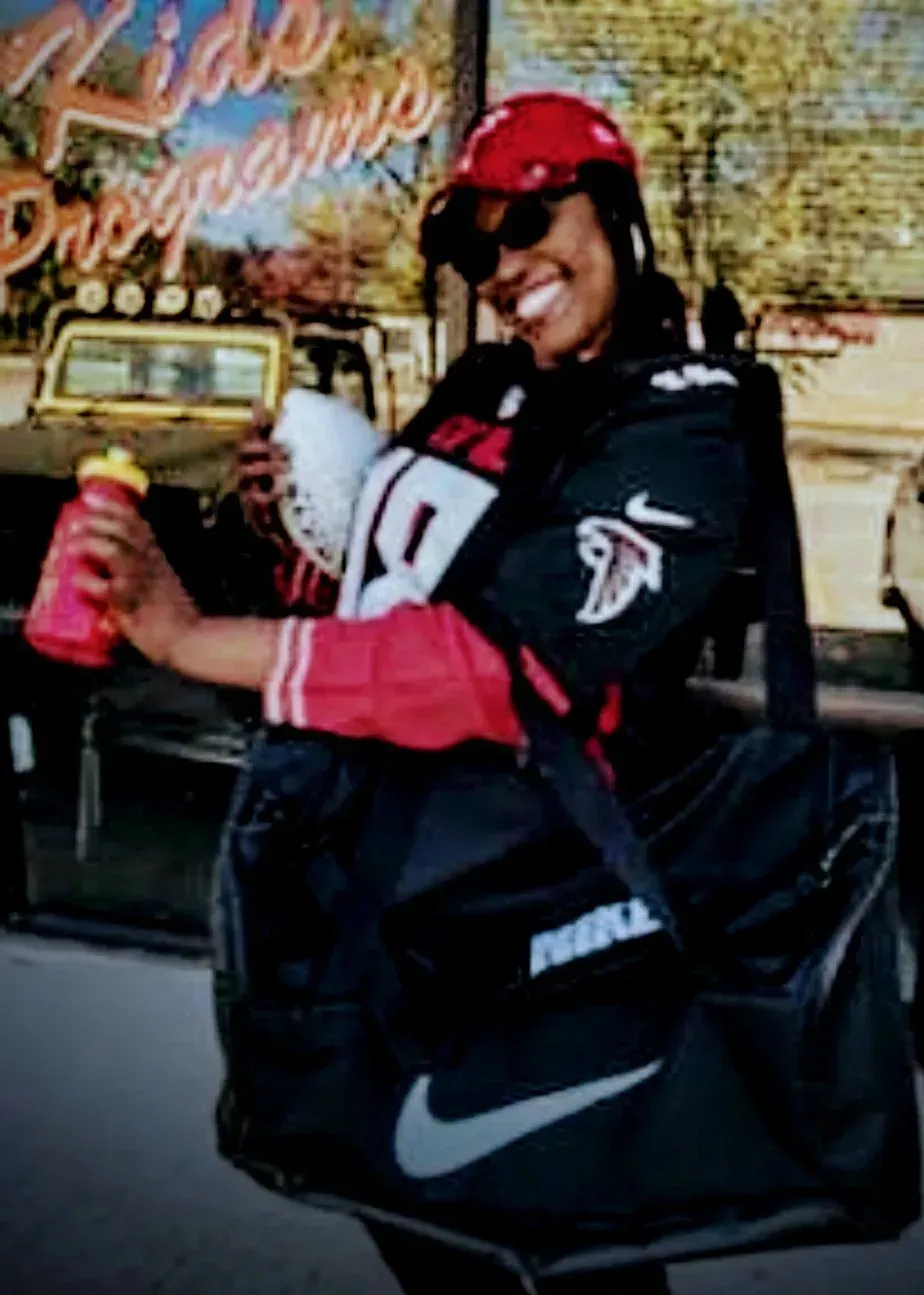 Person in a red cap and black jacket holding a drink and smiling in front of a storefront.