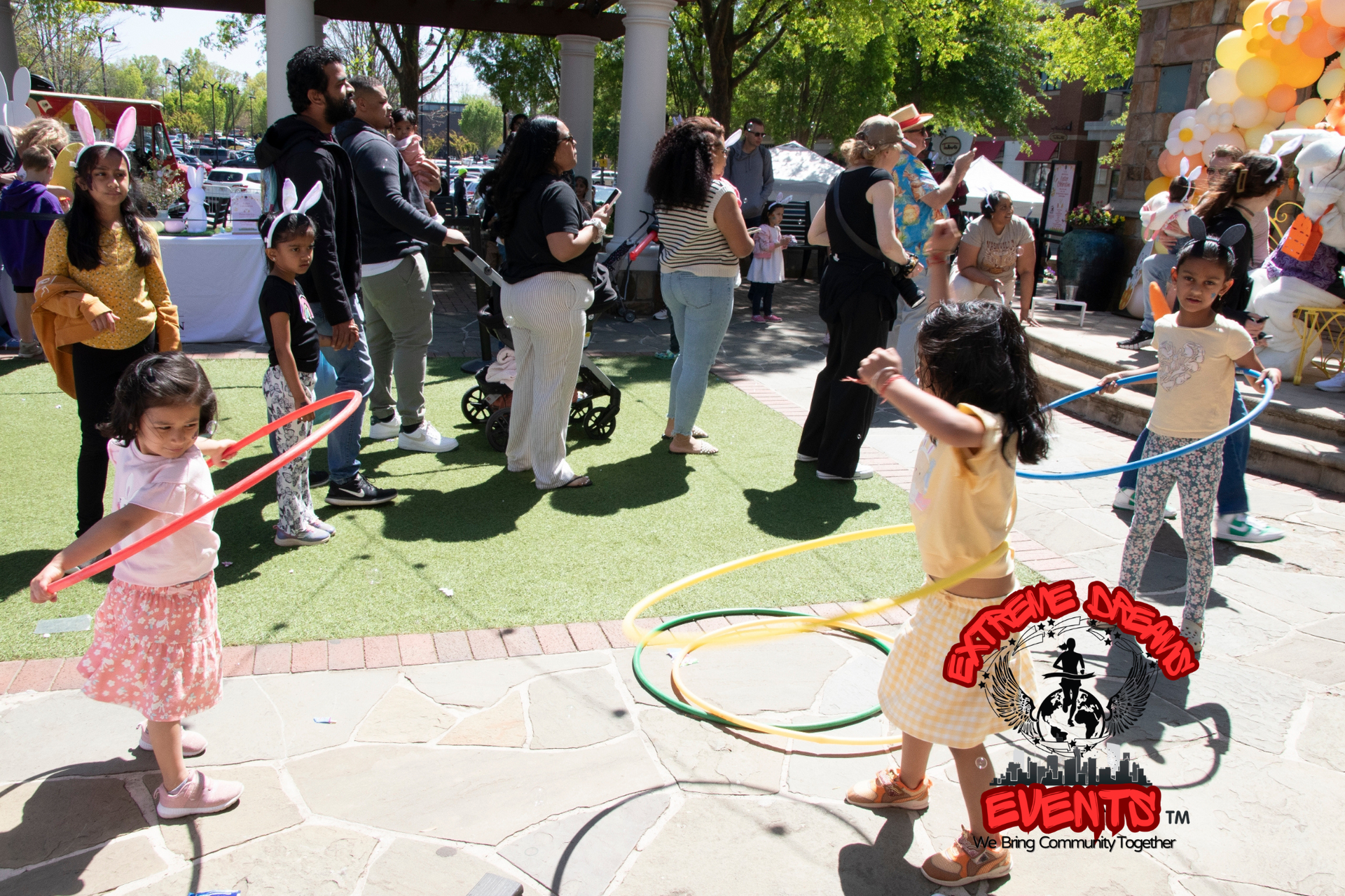 Children hula-hoop at an outdoor event, with adults watching nearby under trees.