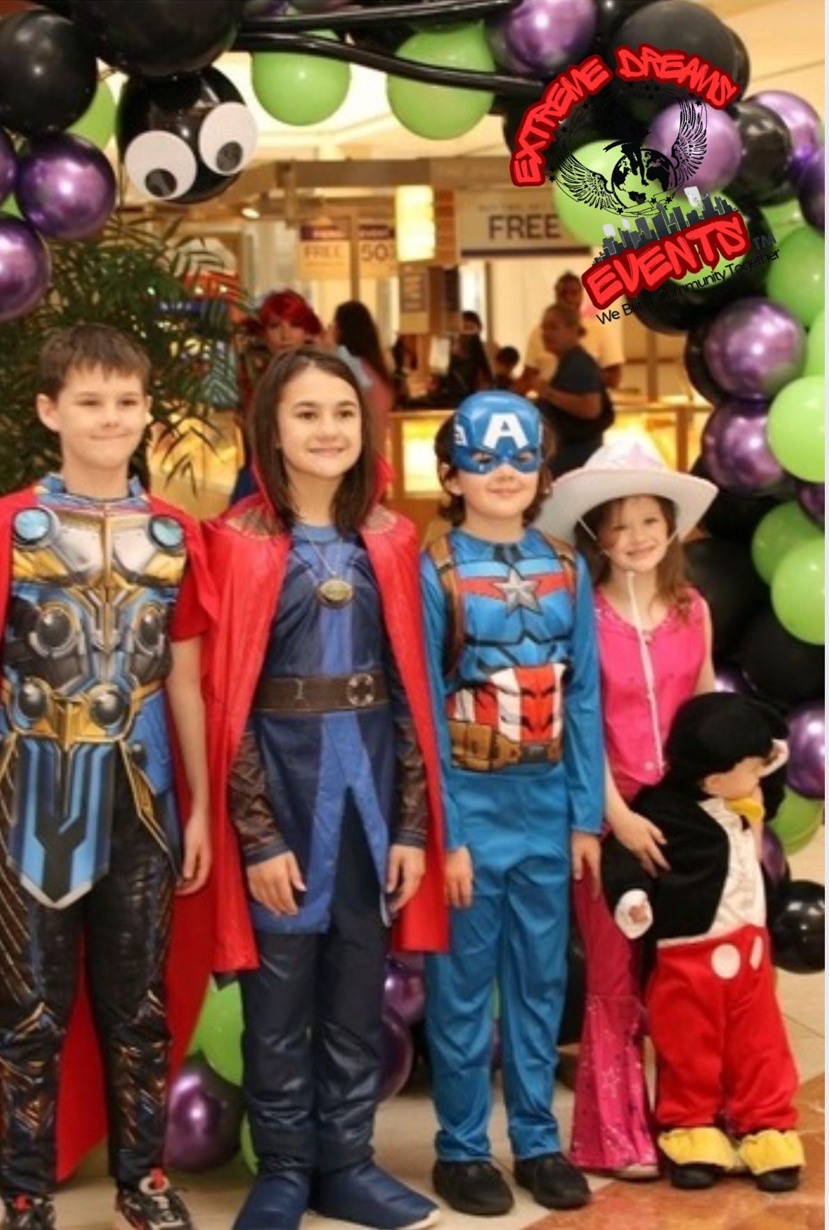 Four children in superhero costumes stand under a balloon arch at a store entrance.