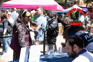 People gathered at an outdoor event booth under a pink canopy, with a wreath and event sign on display.