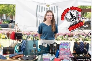 Vendor smiling behind a table of colorful bags and accessories at an outdoor market.