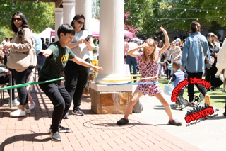 People pulling on a rope outdoors near a brick walkway and white column at a lively event