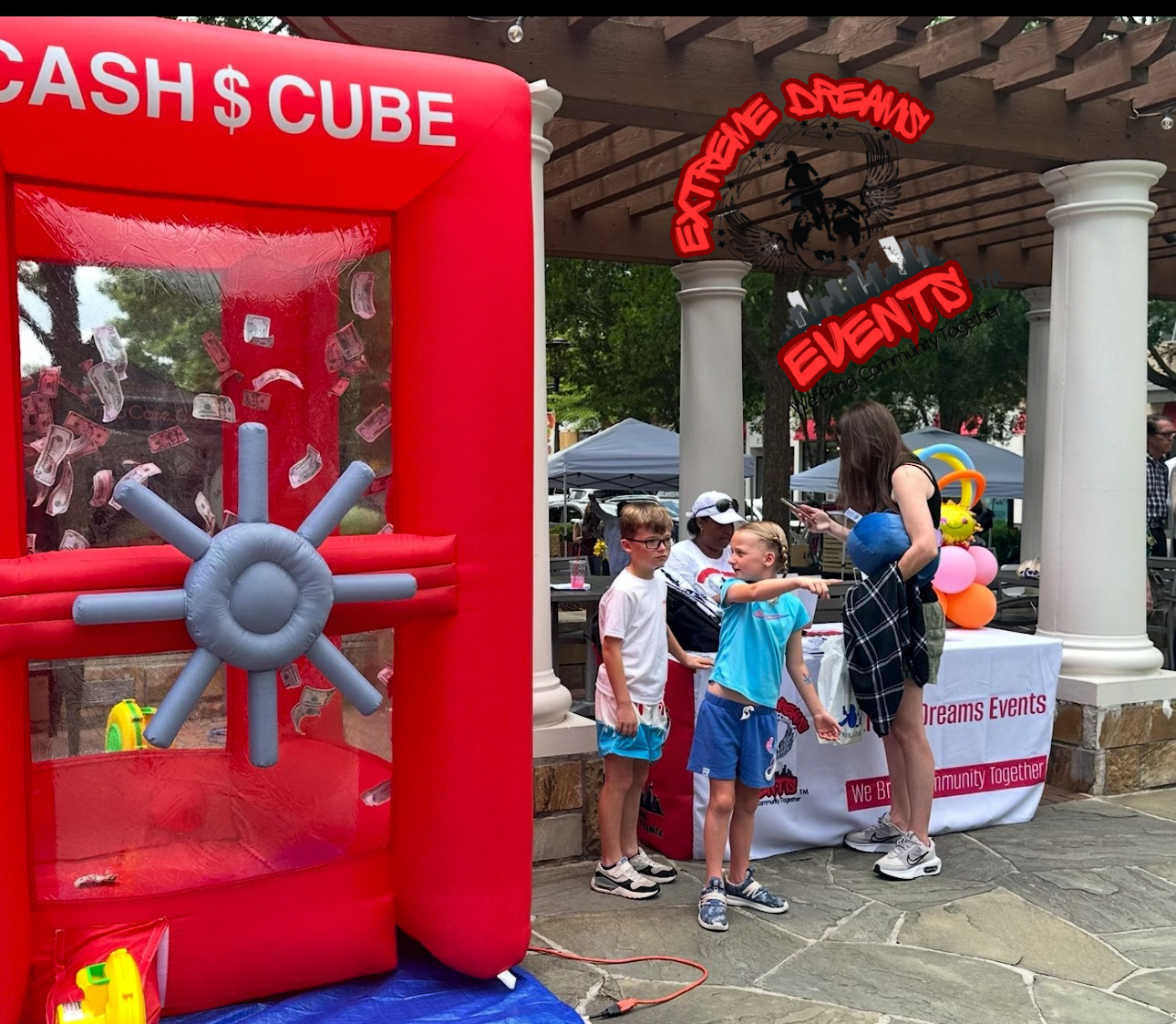 Children stand near a red cash cube inflatable at a community event booth outdoors