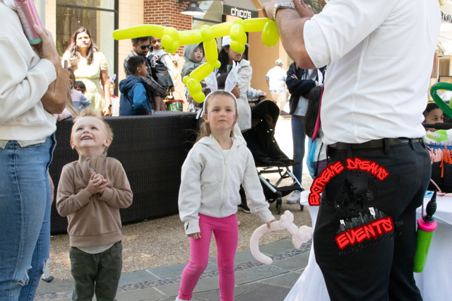 Children watch a street performer with bright yellow balloon animals in a busy outdoor plaza.
