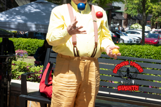 Clown in yellow shirt and red suspenders beside a bench at an outdoor event