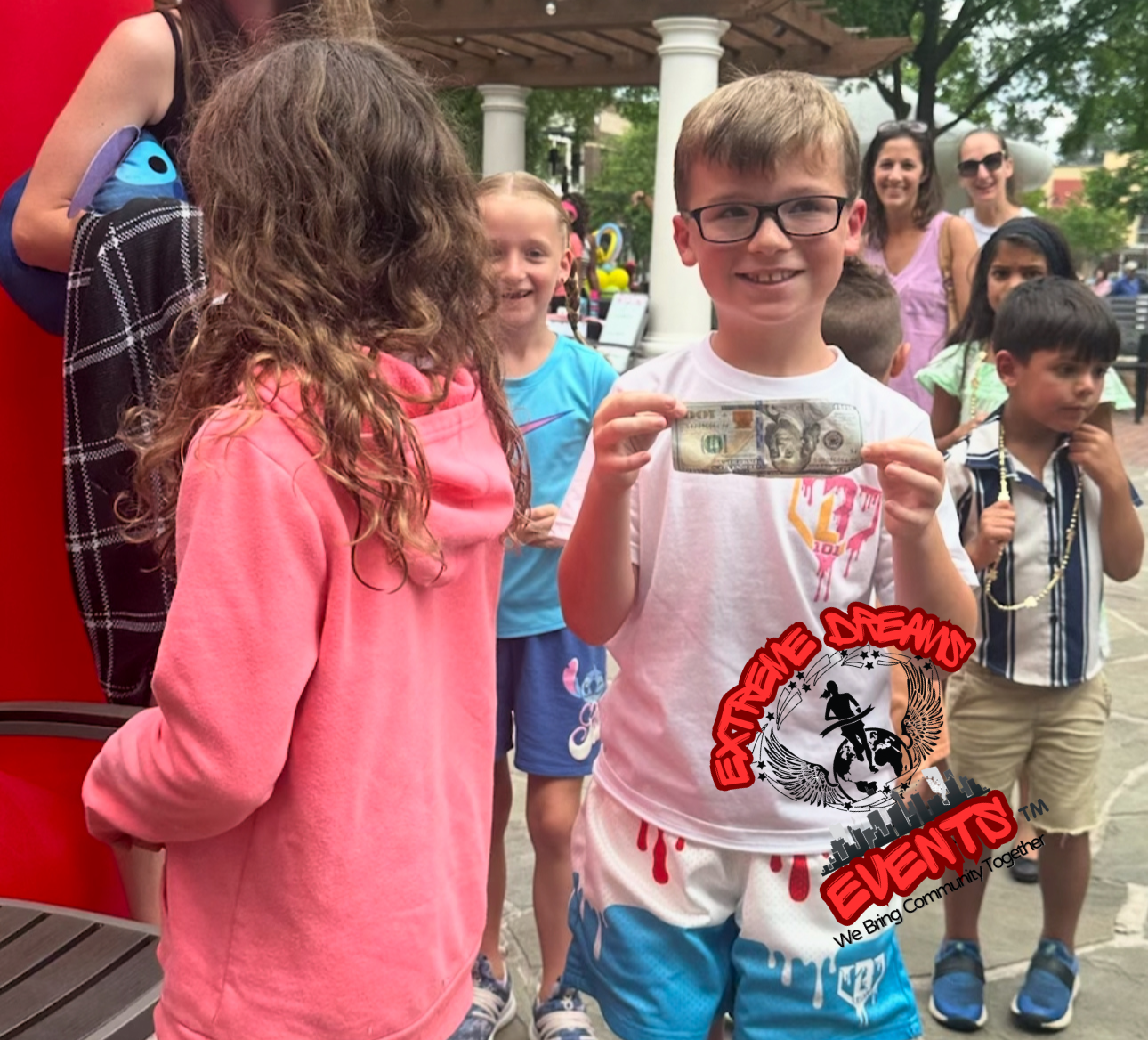 Child smiling and holding a fish at an outdoor event, with other children and adults nearby