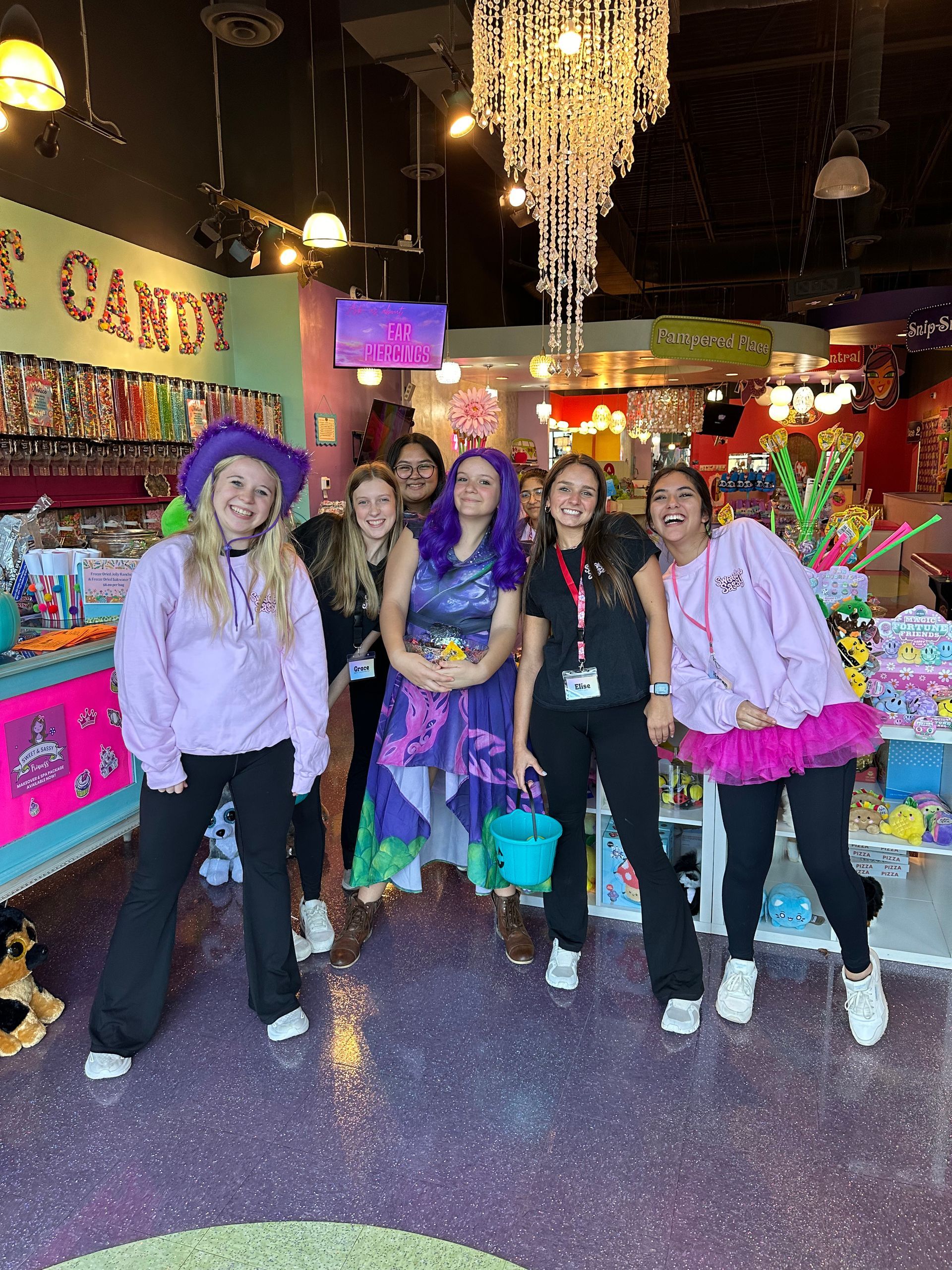 Five people posing in a colorful, decorated indoor shop with sparkling lights and bright displays