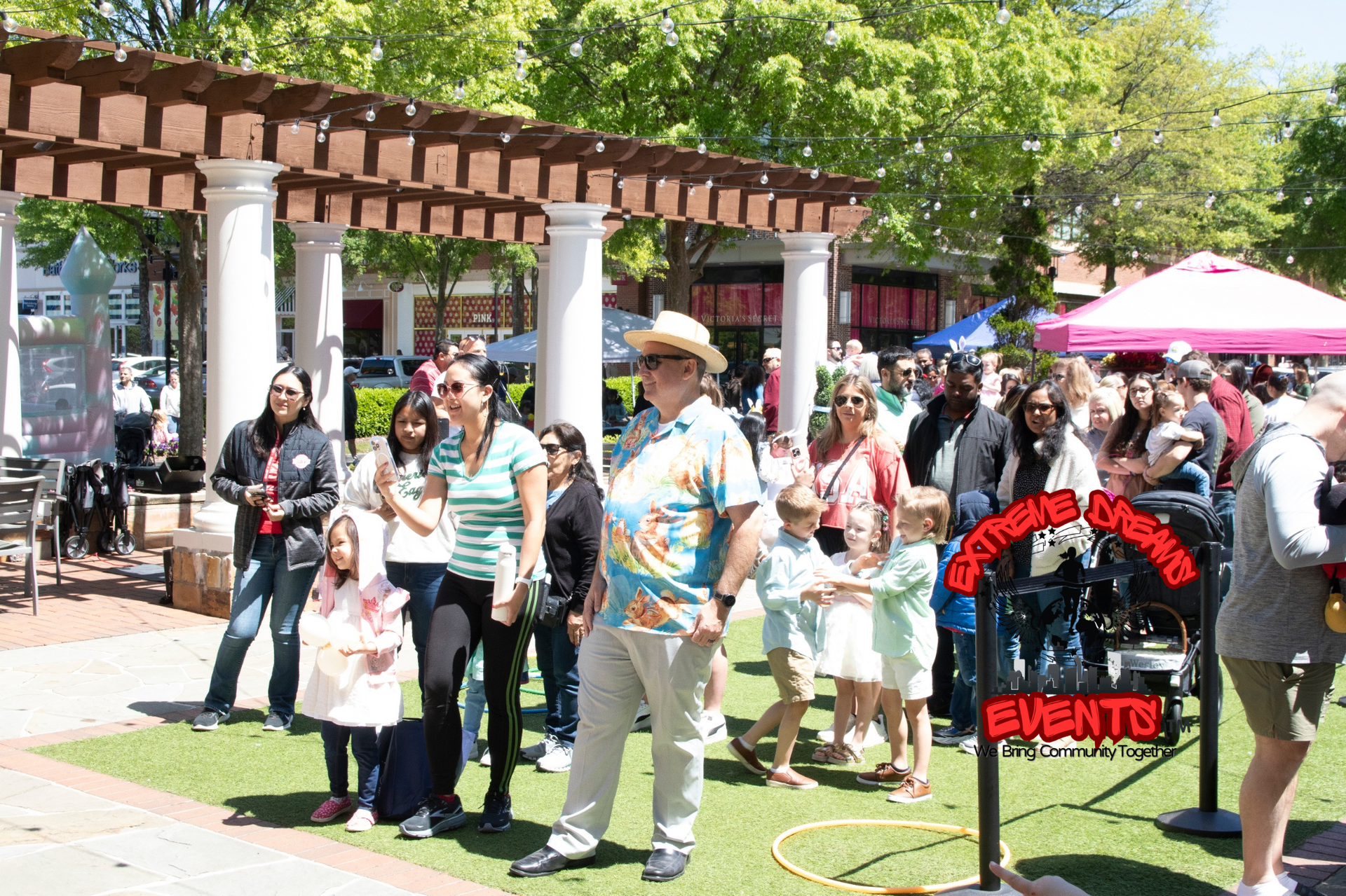 Crowd gathered outdoors near a pergola at a lively event, with a pink vendor tent and people walking around