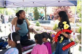 People gathered under a tent at an outdoor event booth, with a woman speaking to children.