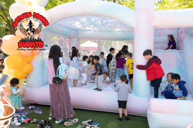 Children playing in a white inflatable bounce house at an outdoor event, with balloons and shoes nearby