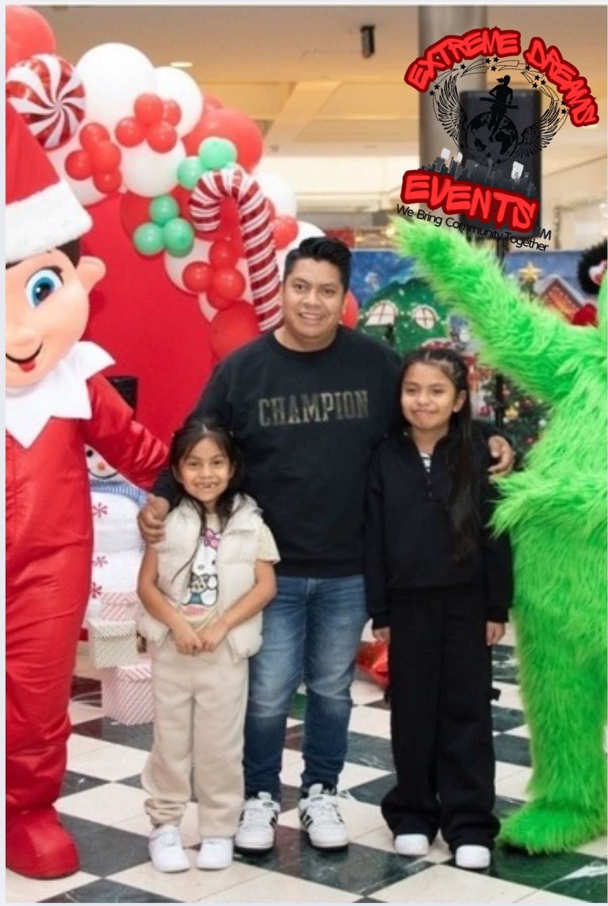 Three people posing with Santa and a green holiday character at a festive event backdrop
