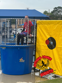 Person seated in a blue dunk tank beside a yellow target panel at an outdoor event