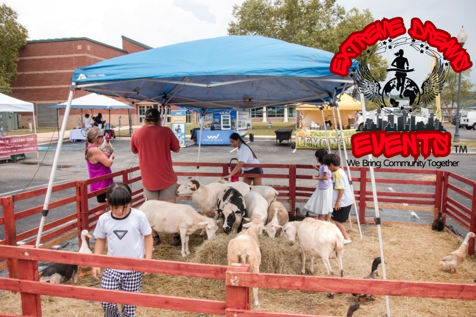 Children petting goats in a red-fenced pen at an outdoor farm event under a blue canopy