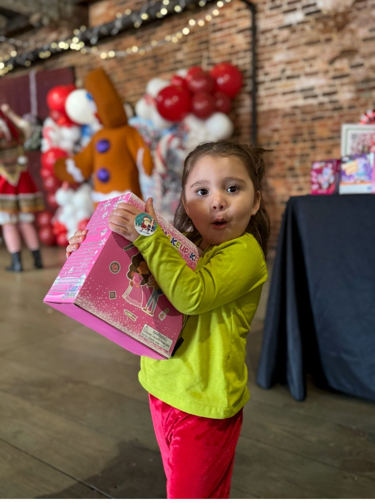 Child holding a pink gift box, standing indoors near balloon decorations and a table