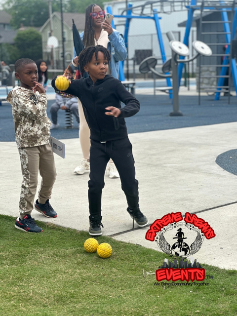 Children playing a tossing game in a park, with yellow balls and playground equipment nearby.