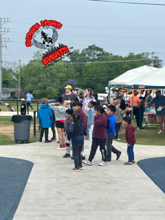 Children standing in line at an outdoor event near a white tent and vendor booth.