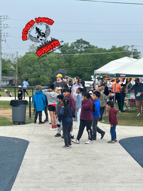 Children walking on a paved path at an outdoor community event with tents and a crowd nearby