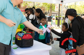 People at an outdoor event exchanging items at a table with backpacks and bags.