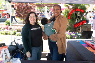 Two people at an outdoor community booth, smiling and holding a small dog, with flowers and tables in the background.