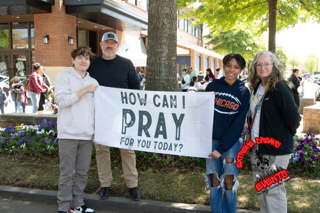 Four people hold a sign reading “HOW CAN I PRAY FOR YOU TODAY?” outdoors near a building.