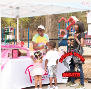 People at an outdoor event booth under a white canopy, with a pink table display and colorful play structures nearby