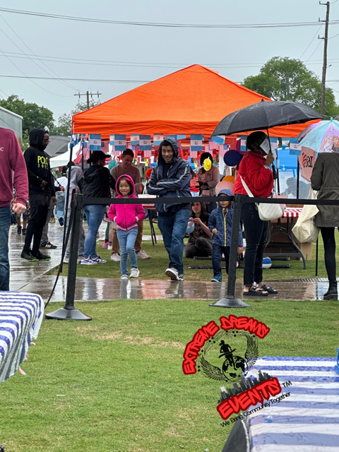 People under an orange tent at a rainy outdoor event, with tables and umbrellas on wet grass.