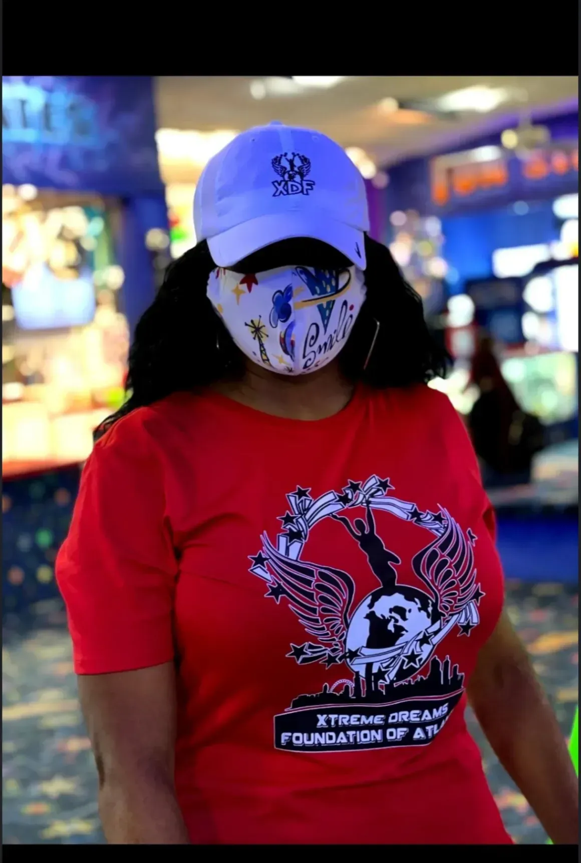 Person in red shirt and white cap with face mask standing in a brightly lit indoor venue.