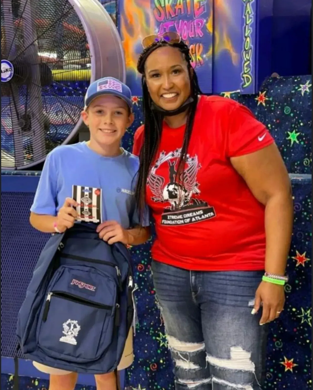 Two people smiling at a skating rink, one holding a backpack, with colorful holiday decorations behind them.