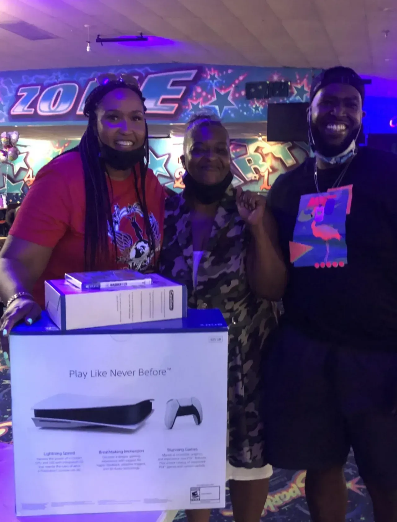 Three people pose indoors with a large boxed gift and a black shopping bag in front of a neon-lit wall.