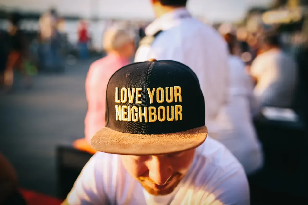 Person wearing a black cap reading “LOVE YOUR NEIGHBOUR” in a crowded outdoor setting