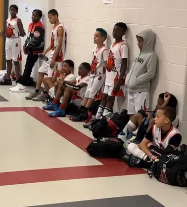 Youth basketball team lined up against a gym wall, some sitting on the floor with bags nearby.