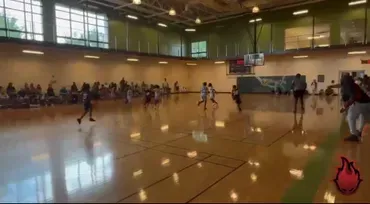 Indoor basketball game in a gym, with players on the court and spectators along the wall.