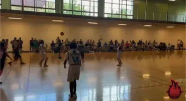Indoor basketball game in a gym, with players on court and spectators seated along the wall.