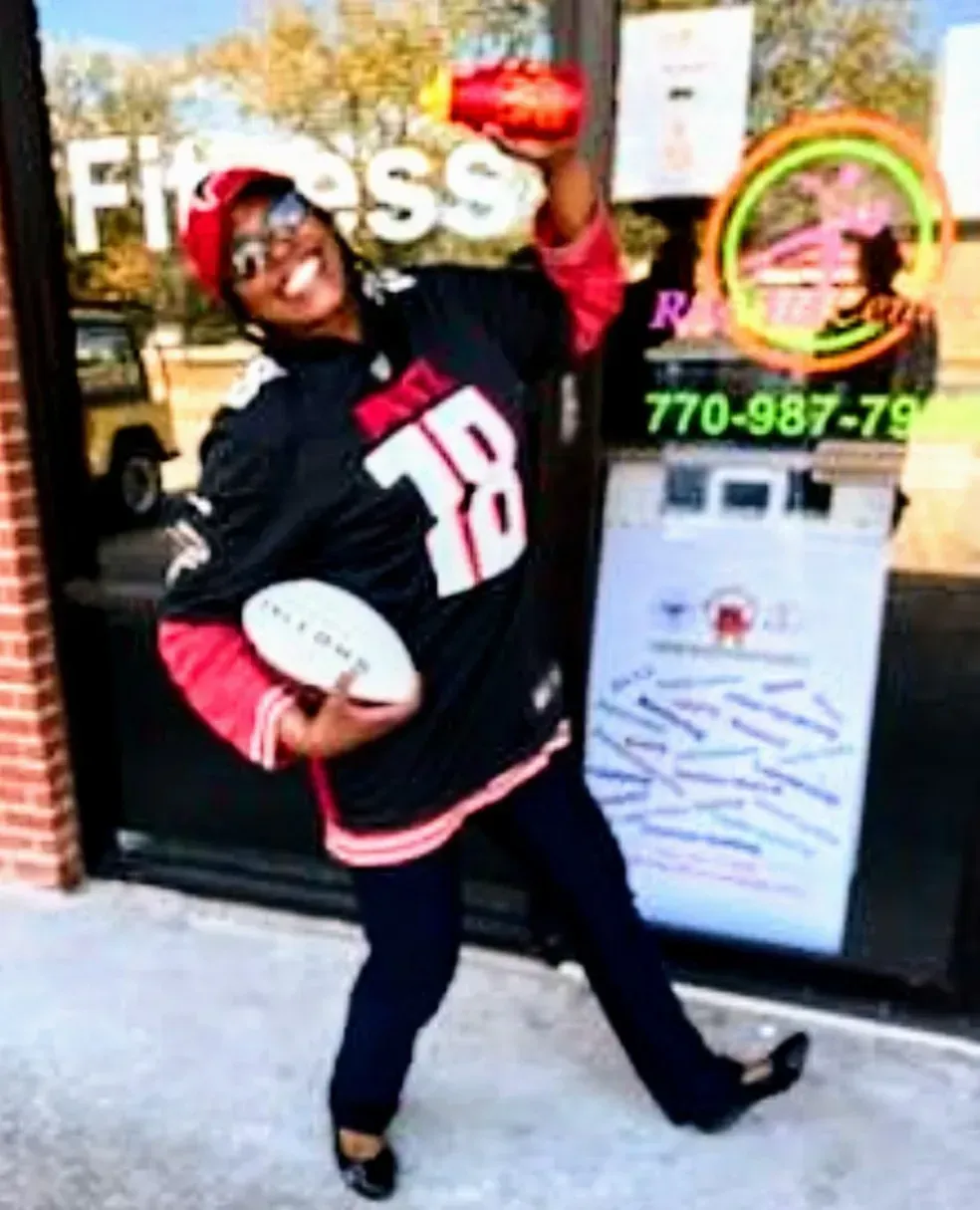 Person in black and pink football jersey posing outside a storefront, holding a football and red helmet.