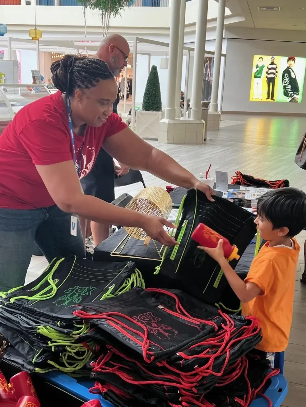 Woman hands black-and-red bags to a child at a booth in a bright indoor hall.